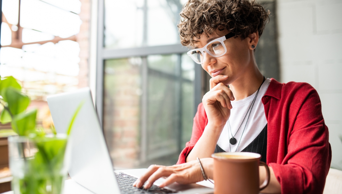 A young woman with curly brown hair and white glasses, wearing a dark pink jacket, ponders the images on her laptop screen, which are not visible to the viewer. In the foreground is a mug and a plant in a glass of water.