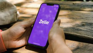 Disembodied hands hold a smartphone featuring the Zelle app outdoors at what appears to be a picnic table.