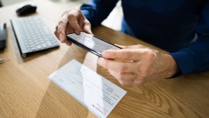 An individual in a dark blue shirt takes a picture of a check with their smartphone; nearby is a wireless keyboard.