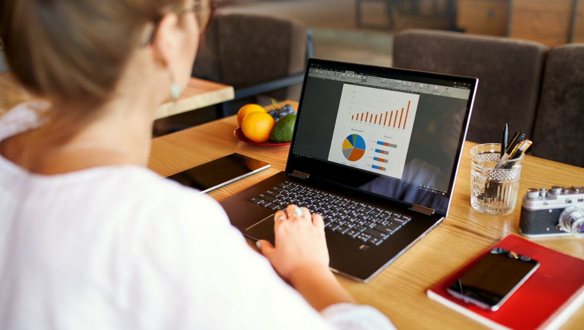A woman works at a wooden table on a laptop displaying colorful charts and graphs, including a bar graph, pie chart, and stacked bar charts. The setting appears to be a casual workspace or café, with a bowl of fruit, a glass of water with pens, a smartphone, a notebook, and a vintage camera on the table.
